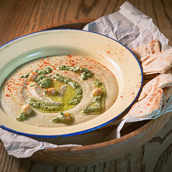 Hummus with green pesto, chickpeas, paprika, olive oil, and pita bread on a wooden table.