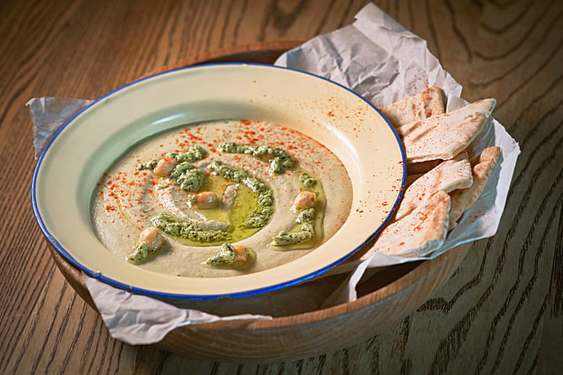 Hummus with green pesto, chickpeas, paprika, olive oil, and pita bread on a wooden table.