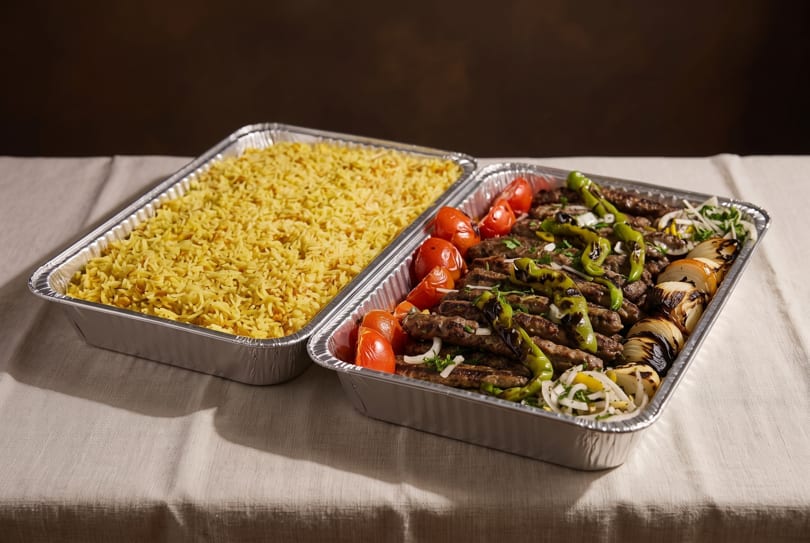 Two catering trays of food, one with yellow rice and the other with grilled meat, vegetables.