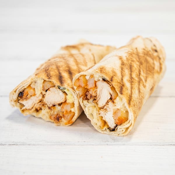 Two grilled chicken wraps with potatoes and sauce, on a white wooden surface.